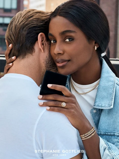 Attractive couple embracing outdoors. The woman, with dark hair, is adorned with layered gold necklaces, bracelets, and a diamond ring.
