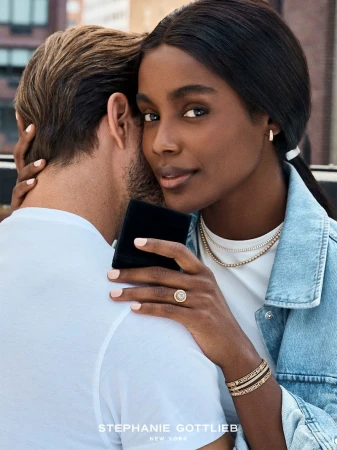 Close-up of a Black woman wearing layered gold jewelry and a denim jacket embracing a man, holding a velvet ring box.