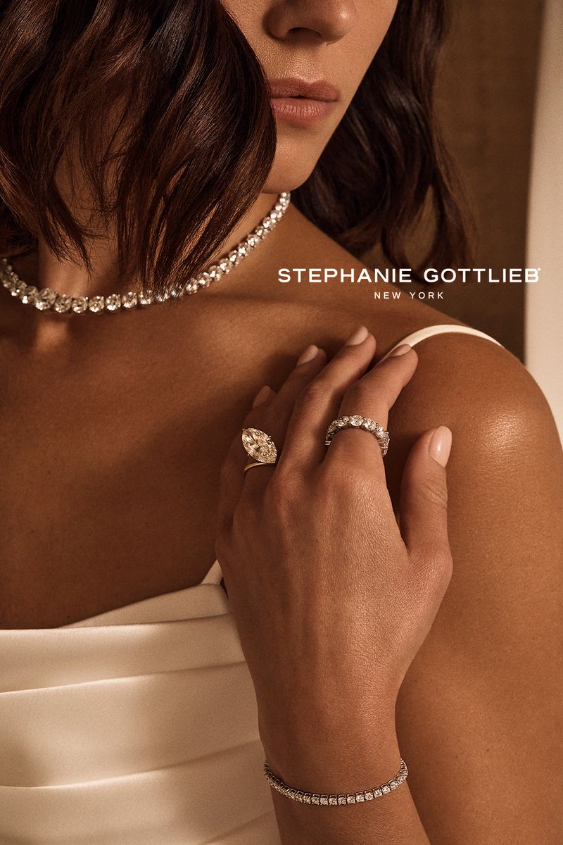 Close-up of a woman showcasing luxury diamond jewelry including a tennis bracelet, necklace, and rings against a neutral backdrop.