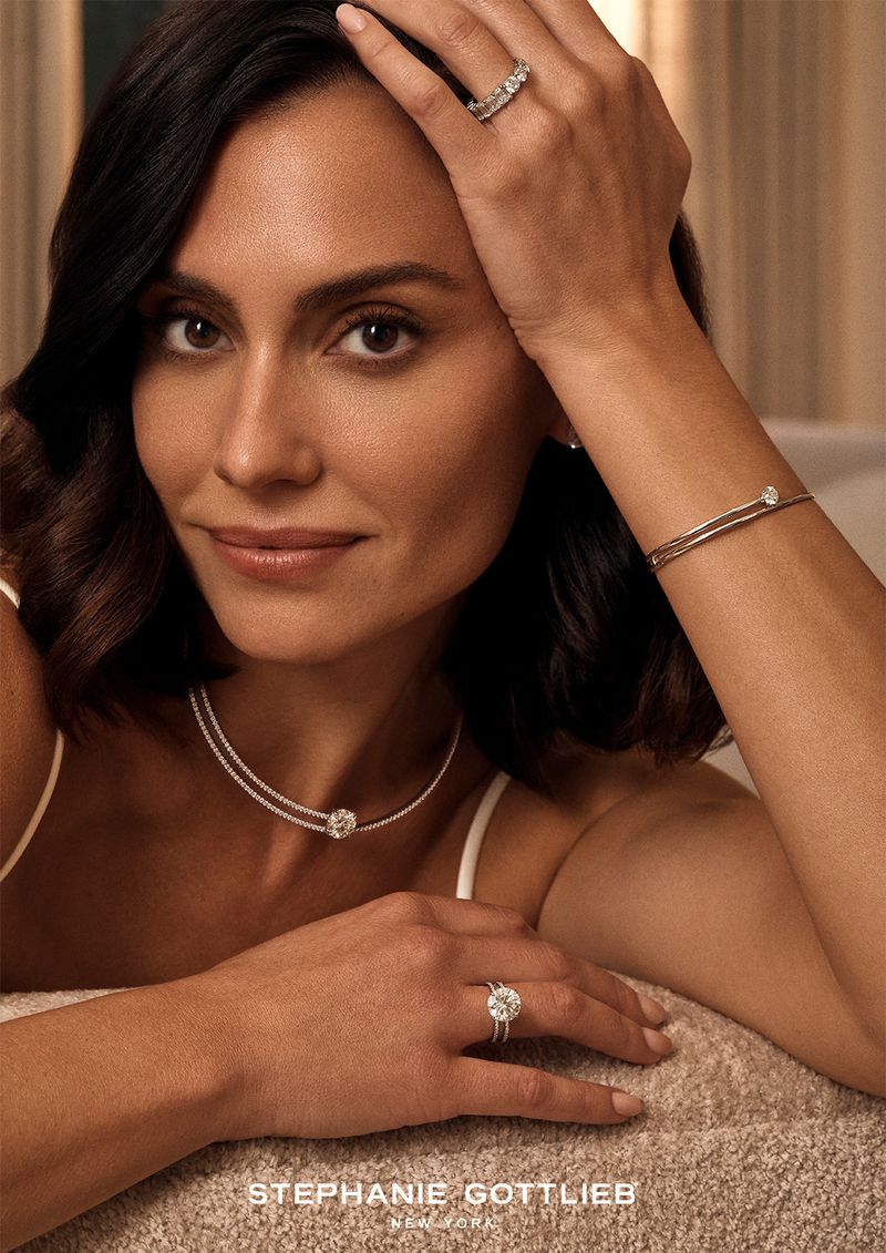 Close-up shot of a woman with dark hair wearing a necklace, bracelet, and rings with gemstones, modelling jewelry in soft lighting.