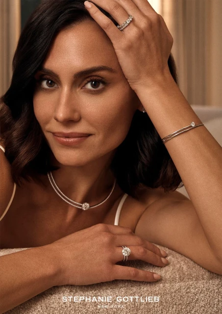 Close-up shot of a woman with dark hair wearing a necklace, bracelet, and rings with gemstones, modelling jewelry in soft lighting.