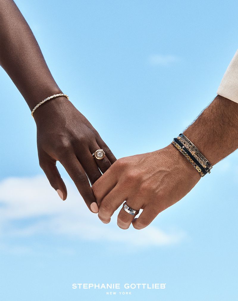 Close-up of an interacial couple holding hands, showcasing gold bracelets and rings against a blurred blue sky background.