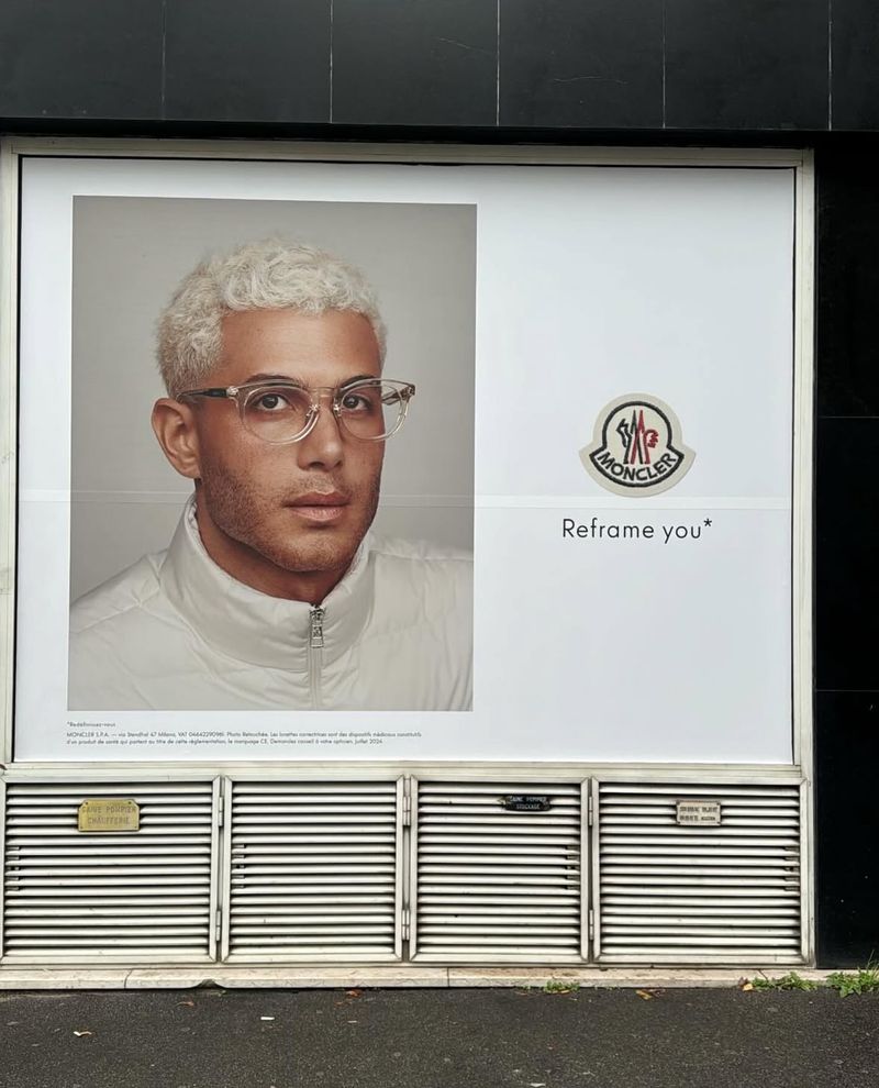Moncler advertisement features a man with platinum blonde hair and glasses wearing a white jacket, next to the Moncler logo.