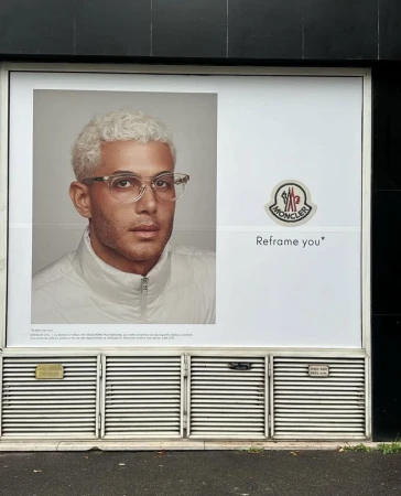 Moncler advertisement features a man with platinum blonde hair and glasses wearing a white jacket, next to the Moncler logo.