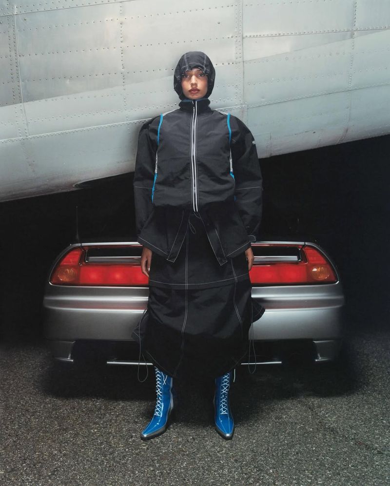 A model in a black jacket and skirt stands in front of a silver sports car and the wing of a silver airplane, creating an automotive fashion shoot.
