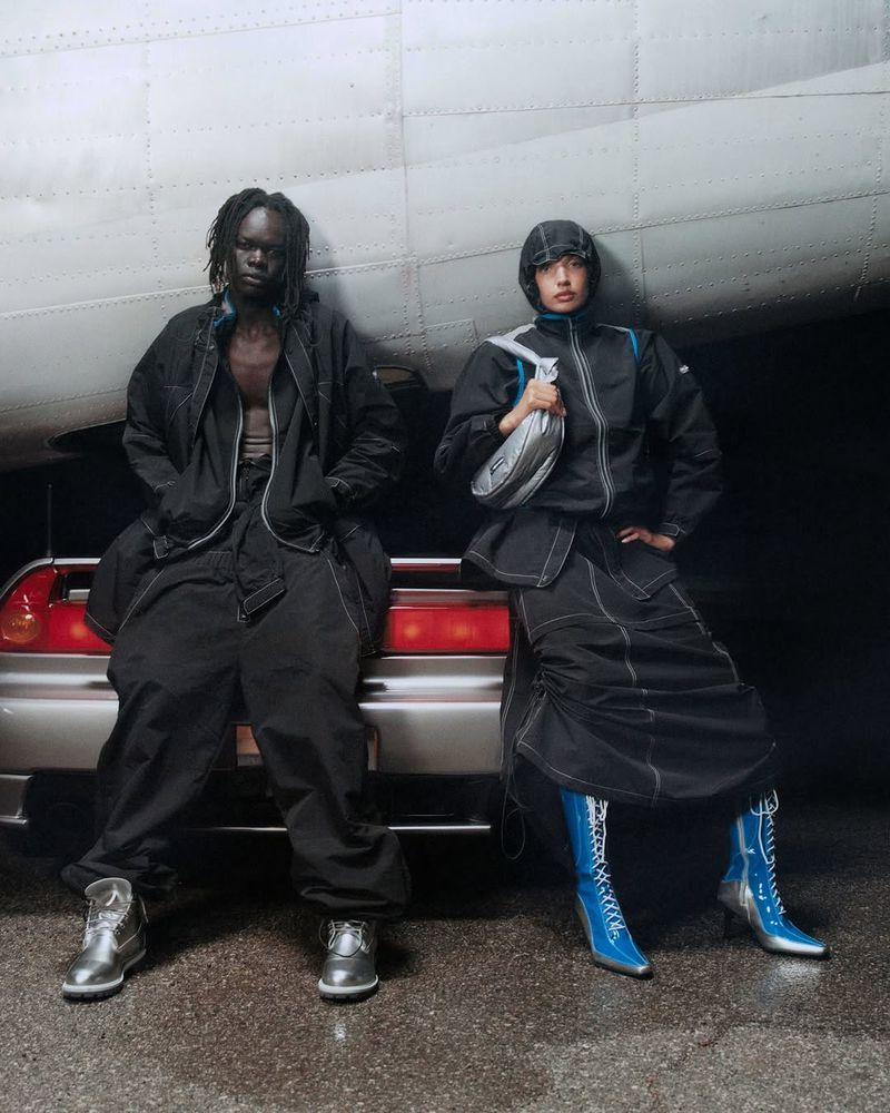 Two models in black and silver clothing pose near a silver car with red tail lights and part of a large airplane fuselage.