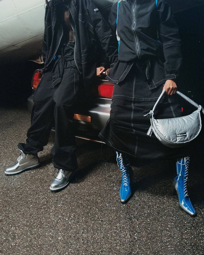 Two people pose in coordinated black outfits with silver shoes and handbag near a silver car and airplane in the background.