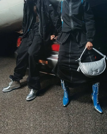 Two people pose in coordinated black outfits with silver shoes and handbag near a silver car and airplane in the background.