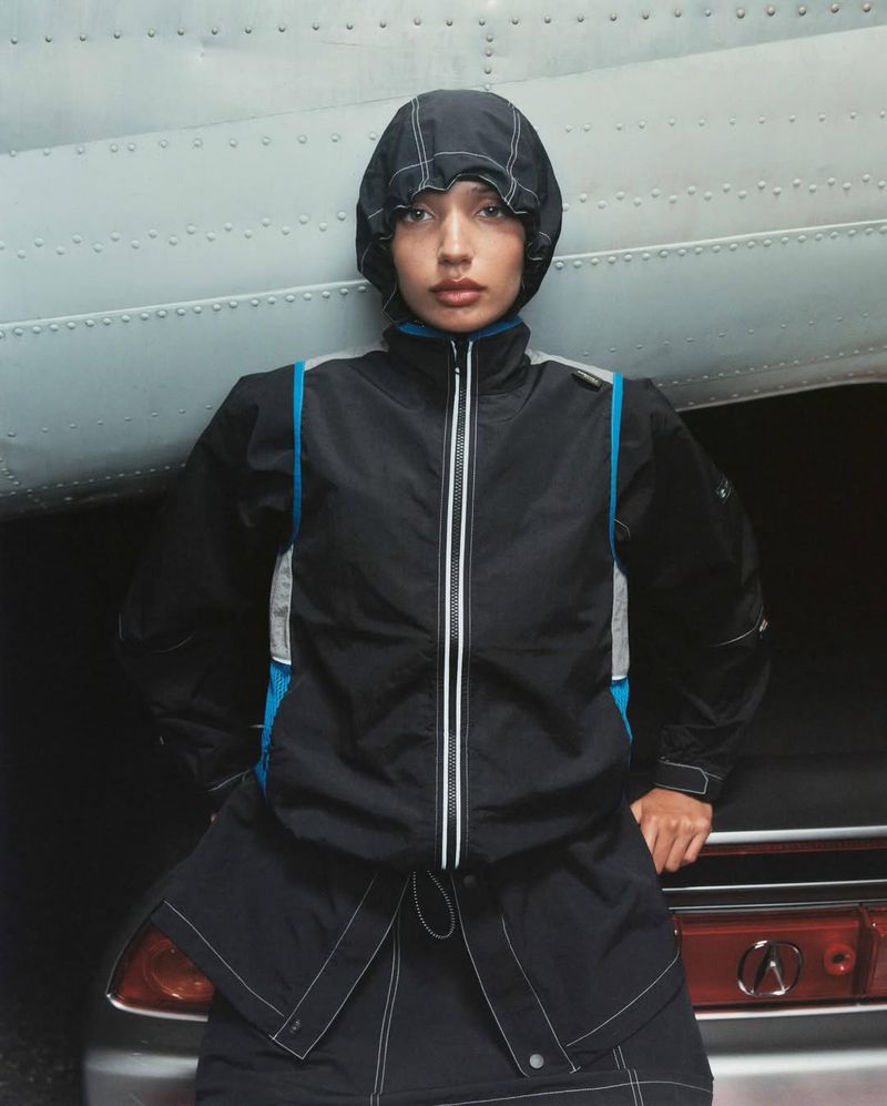 A woman in a black jacket with a hood poses in front of the tail of a silver airplane; studio shot.