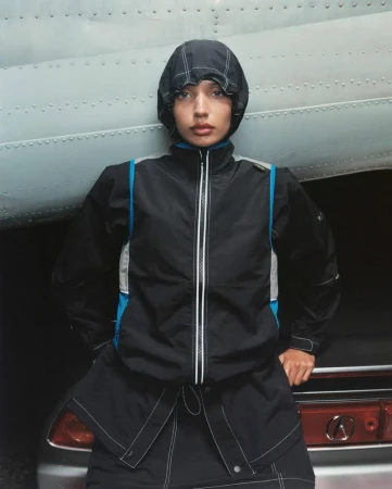 A woman in a black jacket with a hood poses in front of the tail of a silver airplane; studio shot.
