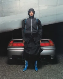 Model standing in black hooded jacket and matching skirt with blue lace-up boots against a car and aircraft exterior