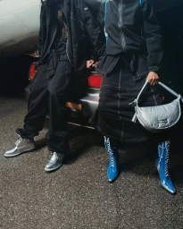 Two models wearing dark technical apparel leaning against the back of a car at night, holding a silver shoulder bag