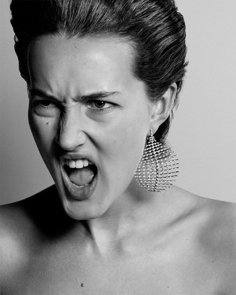 Striking black and white portrait of an adult woman shouting with an expressive face, statement earrings, and bare shoulders against a plain backdrop.