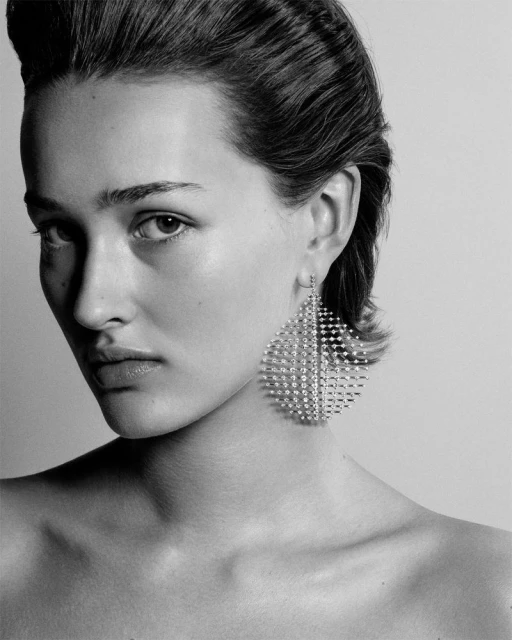 Monochrome portrait of a female model with sleek hair, wearing large geometric earrings against a plain grey background in a studio shot.