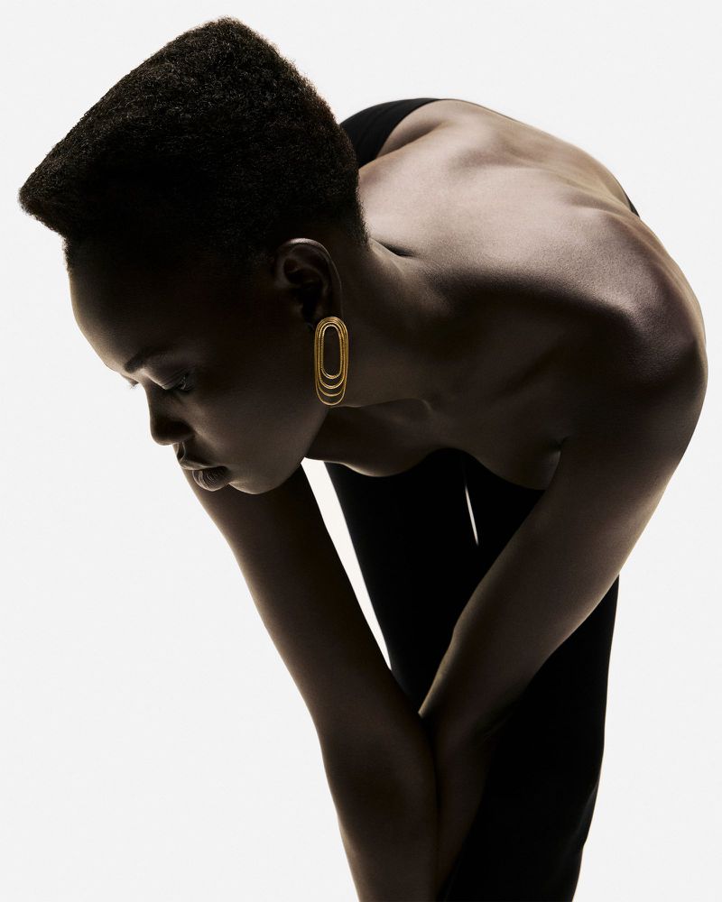 Elegant studio shot of a slender Black woman with gold earrings bending forward, showcasing her bare back and a minimalist fashion aesthetic.