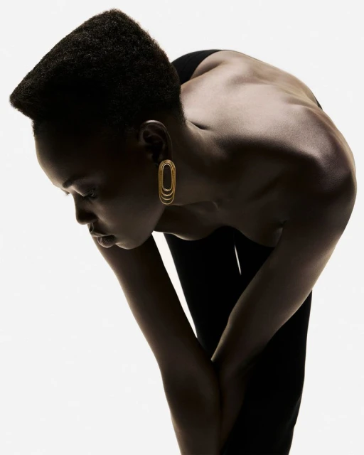 Elegant studio shot of a slender Black woman with gold earrings bending forward, showcasing her bare back and a minimalist fashion aesthetic.