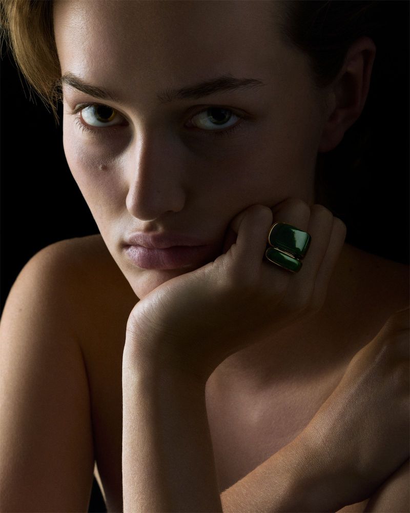 Portrait of a woman with fair skin and short blonde hair, wearing a large, square, emerald green ring, resting her face on her hand.