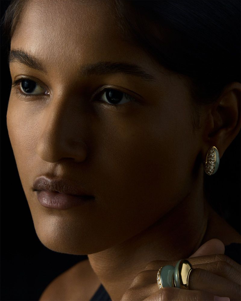 Close-up portrait of a woman with brown skin wearing oval-shaped earrings and a ring with gold and green stones against a black background.