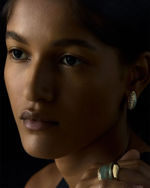 Close-up portrait of a woman with brown skin wearing oval-shaped earrings and a ring with gold and green stones against a black background.