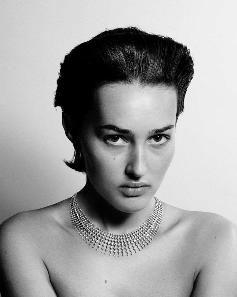 Close-up portrait of an adult woman with short dark hair and light skin wearing a diamond necklace against a white background.