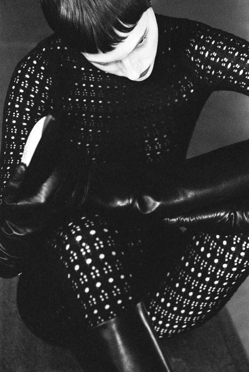 Monochrome fashion portrait of a person in a patterned top pulling on a black, shiny boot, indoors, against a neutral background.