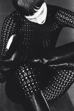 Monochrome fashion portrait of a person in a patterned top pulling on a black, shiny boot, indoors, against a neutral background.