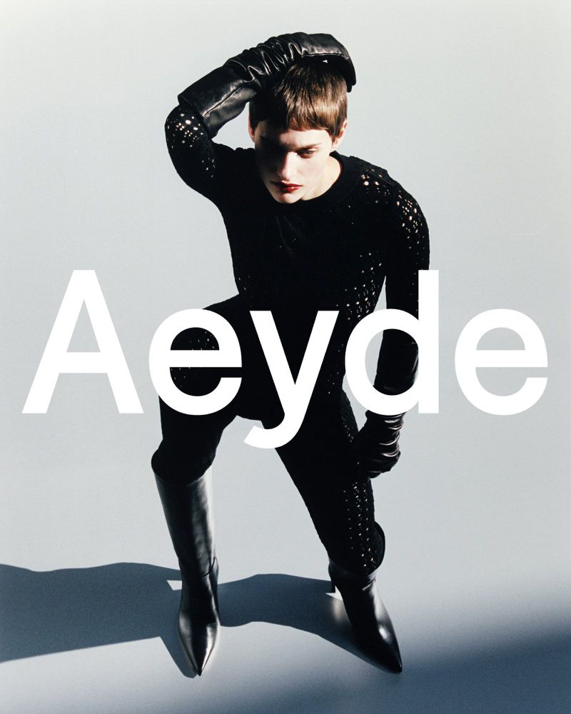 A person poses in black clothing and boots against a neutral background. The model is wearing gloves and looks down in a fashion shot.
