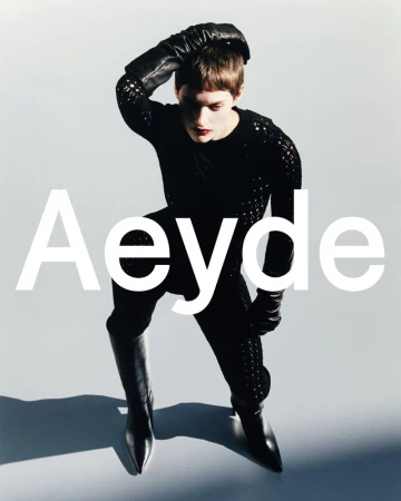 Model in black textured top, leather gloves and boots posing against a grey seamless background for Aeyde