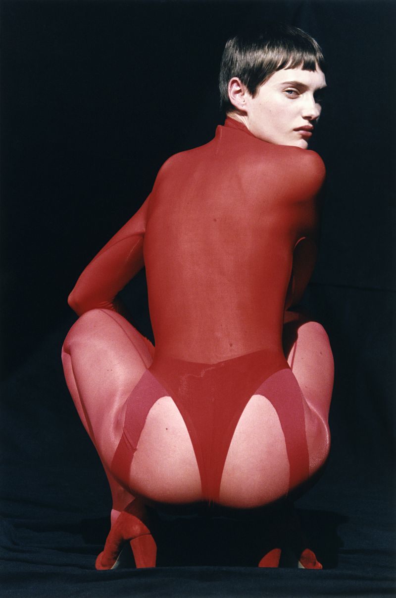 Person crouching in a red bodysuit looking over their shoulder against a dark backdrop in studio lighting, fashion and editorial shot.