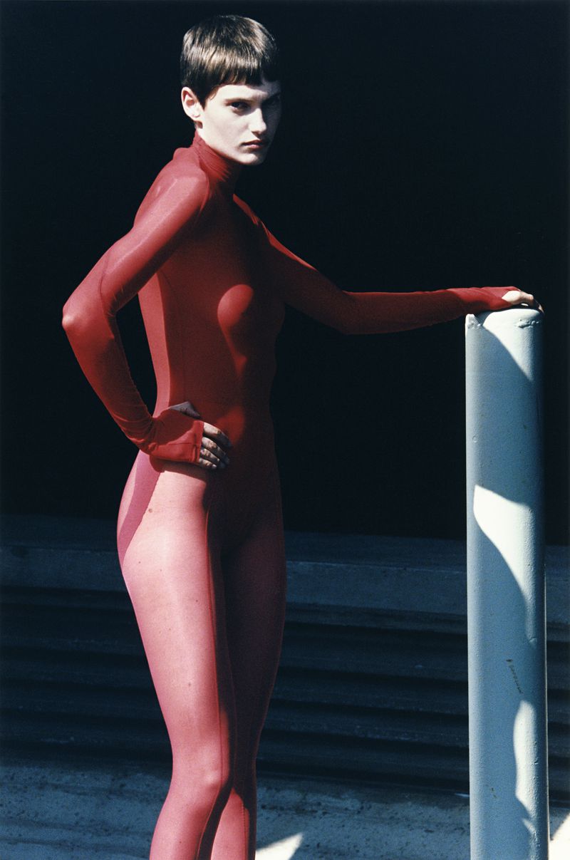 A woman in a full red bodysuit poses next to a concrete pillar against a dark background with visible concrete steps in shadow.
