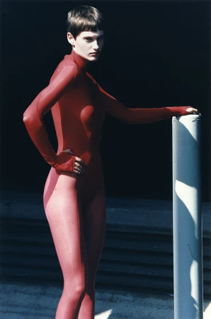 A woman in a full red bodysuit poses next to a concrete pillar against a dark background with visible concrete steps in shadow.