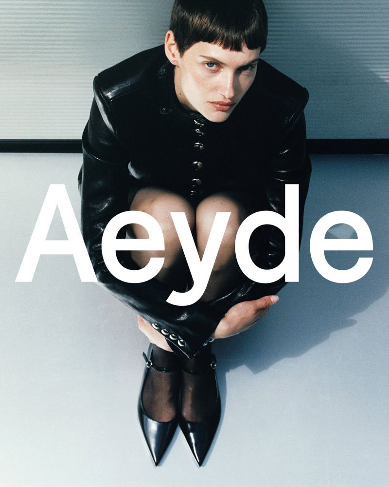 Woman in black jacket and Mary Jane heels sits against a minimalist wall with the logo 'Aeyde' in bold white text.