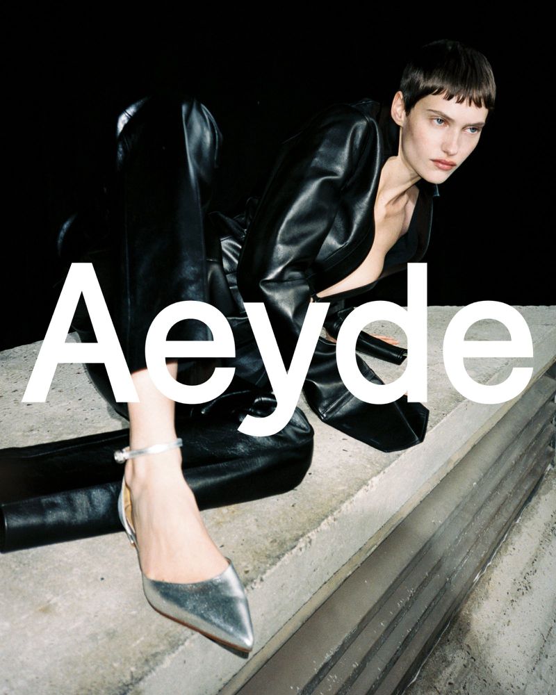 A woman with short hair is wearing a black leather jacket and pants with silver shoes. The brand name Aeyde is in white letters.