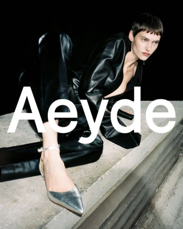 A woman with short hair is wearing a black leather jacket and pants with silver shoes. The brand name Aeyde is in white letters.