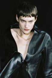 Model with short dark hair wearing a black leather jacket against a dark background for Aeyde Pre-Fall 24.
