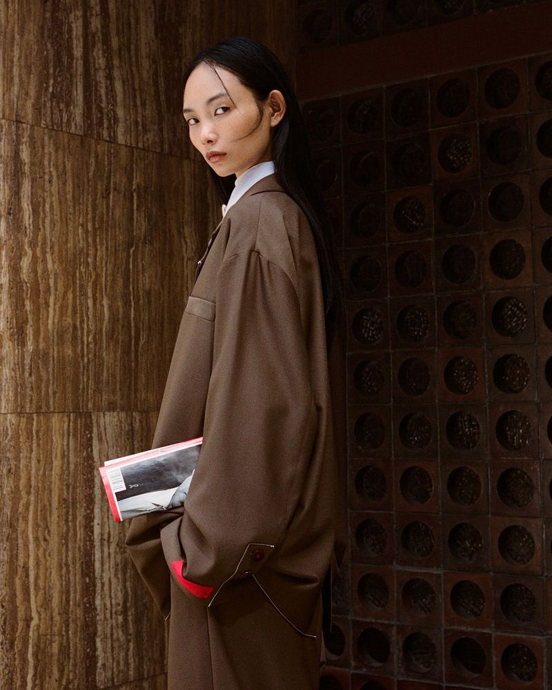 A portrait of an Asian model in an oversized brown suit, holding books and looking at the camera, shot against a textured wall.
