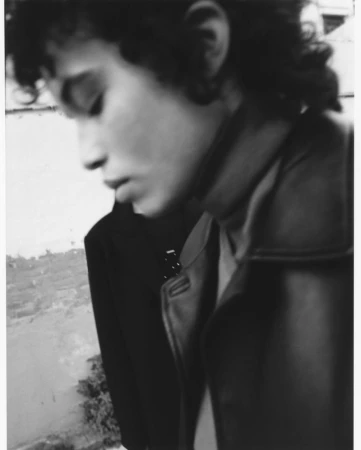 Monochrome portrait of a young adult with curly hair wearing a jacket and dark shirt against an abstract textural background.