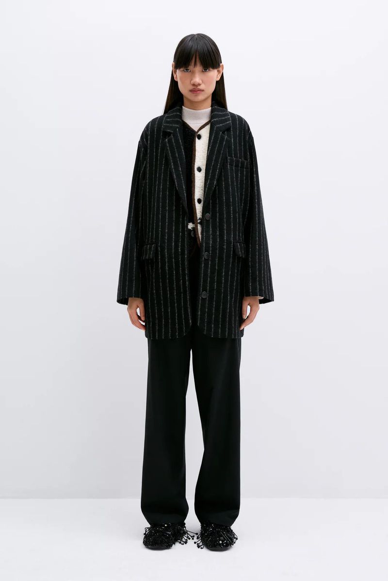 Standing Asian woman in oversized pinstripe blazer, neutral top, and wide-leg pants against a plain white backdrop in a fashion-forward pose.