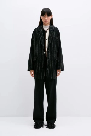 Standing Asian woman in oversized pinstripe blazer, neutral top, and wide-leg pants against a plain white backdrop in a fashion-forward pose.