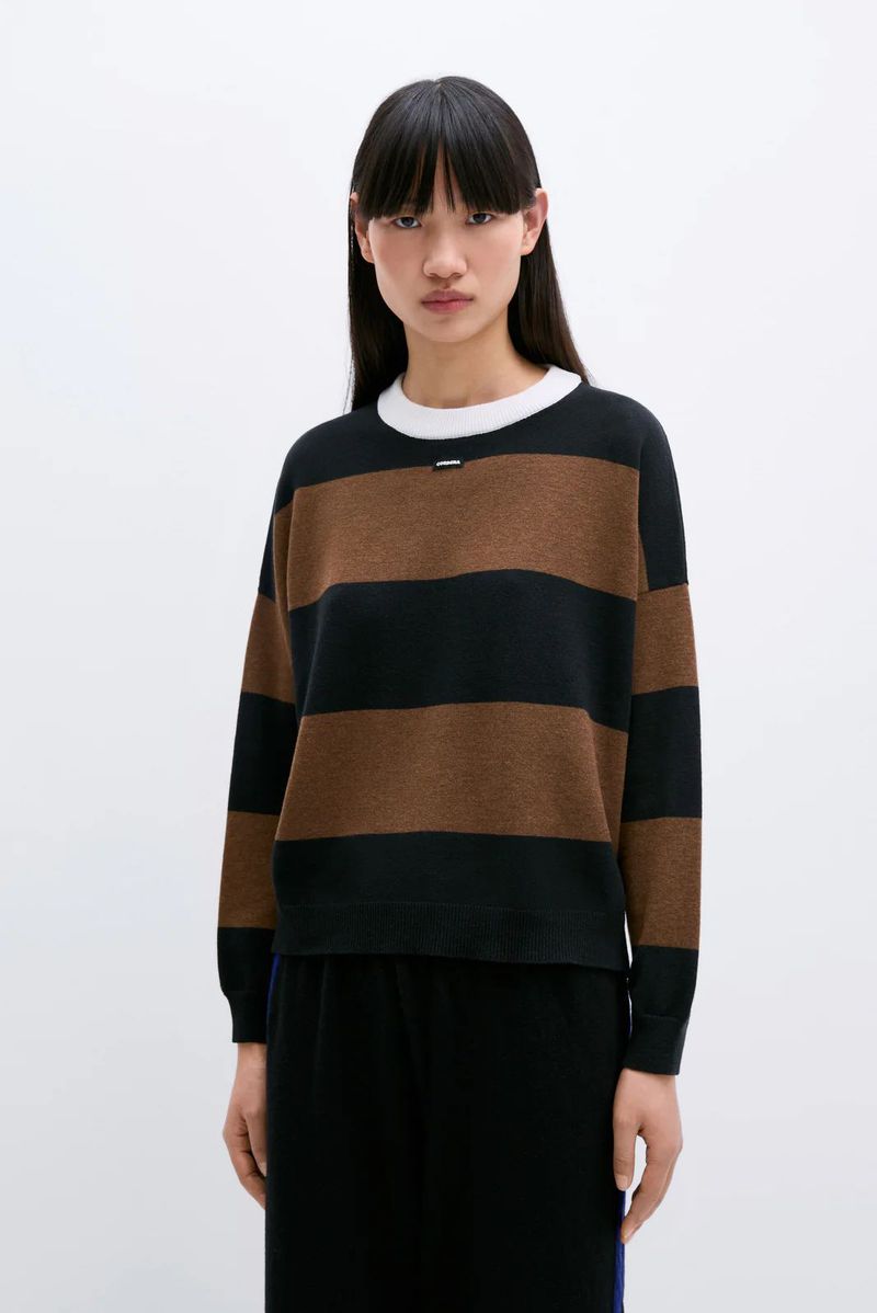 A woman with dark hair wears a horizontally striped brown and black long-sleeved shirt in a studio setting, gazing directly at the camera.