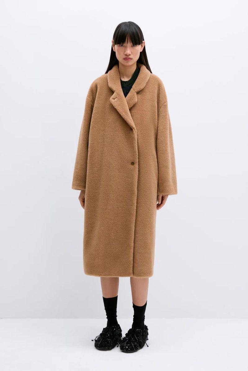 Woman in a tan teddy coat standing in front of a white background. She has long dark hair and is wearing black socks and slippers.