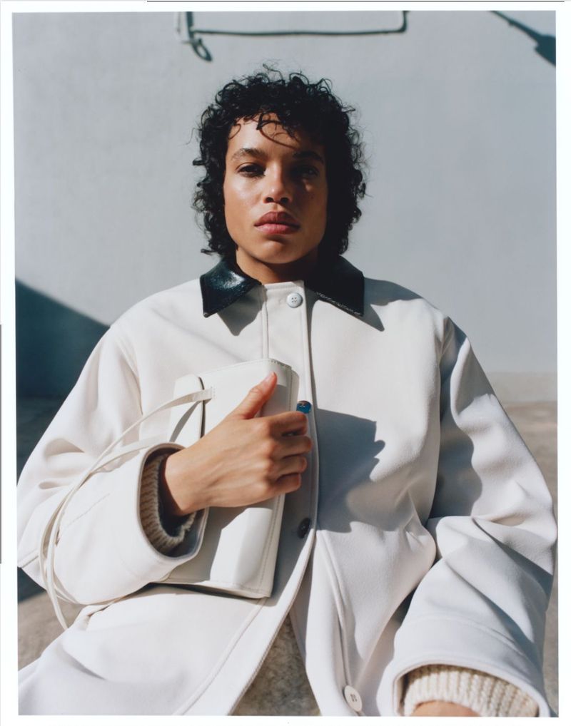 Woman in a cream-colored jacket with a dark collar holds a matching purse, posing against a neutral wall in a fashion editorial shot.