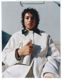 Model with dark curly hair wearing an off-white jacket with a dark collar holding a white handbag
