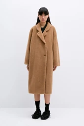 Model standing against a plain white background wearing a long brown textured coat and black shoes