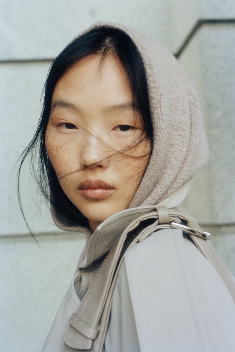Close-up of a young adult Asian woman with freckles and dark hair, wearing a light brown headscarf and coat with a shoulder bag.