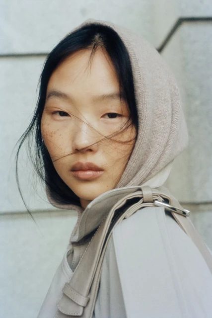 Close-up of a young adult Asian woman with freckles and dark hair, wearing a light brown headscarf and coat with a shoulder bag.