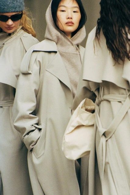 Three women stand in a row wearing trench coats and hoodies. The central woman has an Asian appearance and light skin.