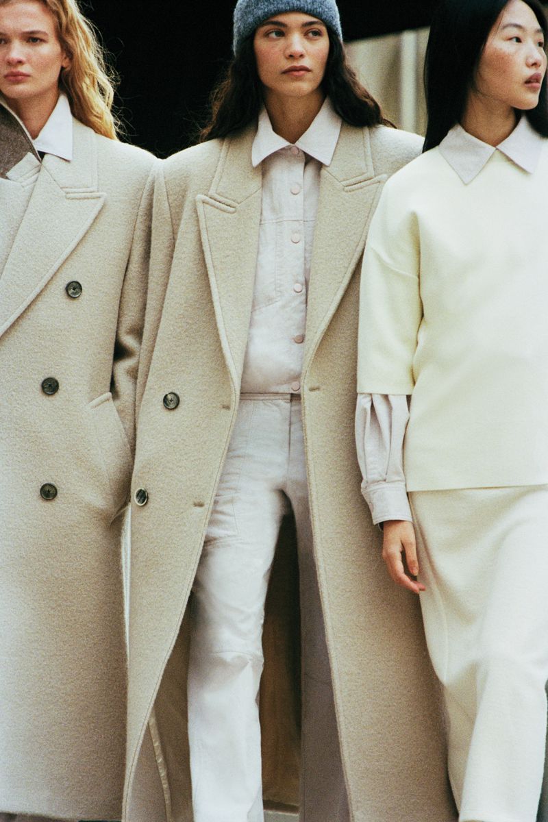 Three women in stylish neutral-toned coats and apparel. The central figure wears a blue beanie, creating a fashionable and eye-catching group shot.