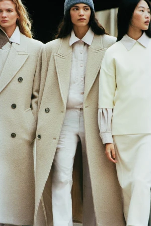 Three women in stylish neutral-toned coats and apparel. The central figure wears a blue beanie, creating a fashionable and eye-catching group shot.