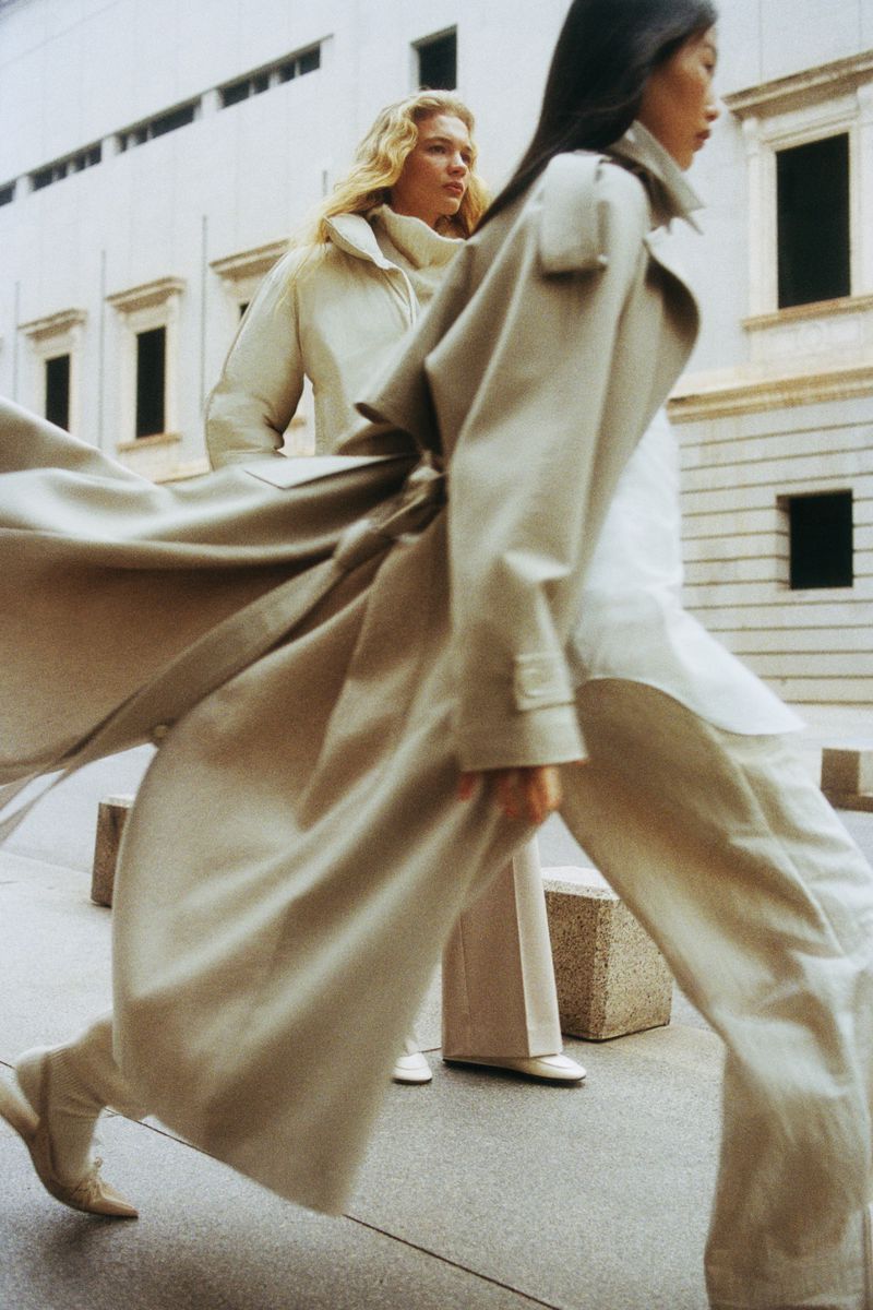 Two women in long coats walk on a city sidewalk with neutral stone buildings. One woman is mid-stride, coat flowing open.